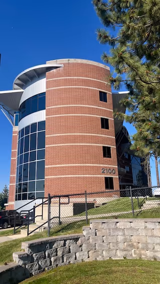 Brick cylindrical building exterior with large glass windows and a stairway on a grassy slope.