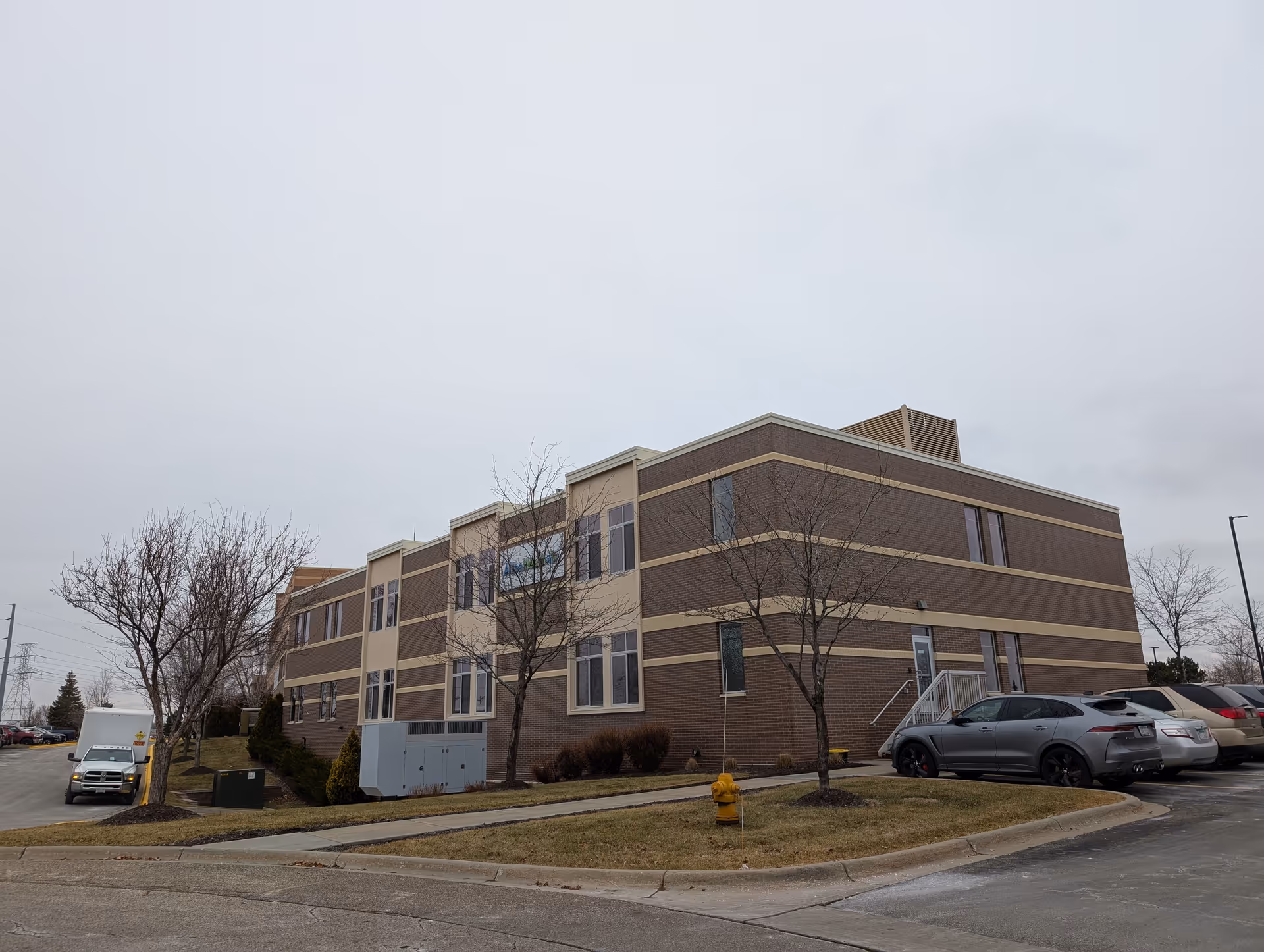 Two-story brick senior living building with parked cars, leafless trees, and a fire hydrant under an overcast sky.