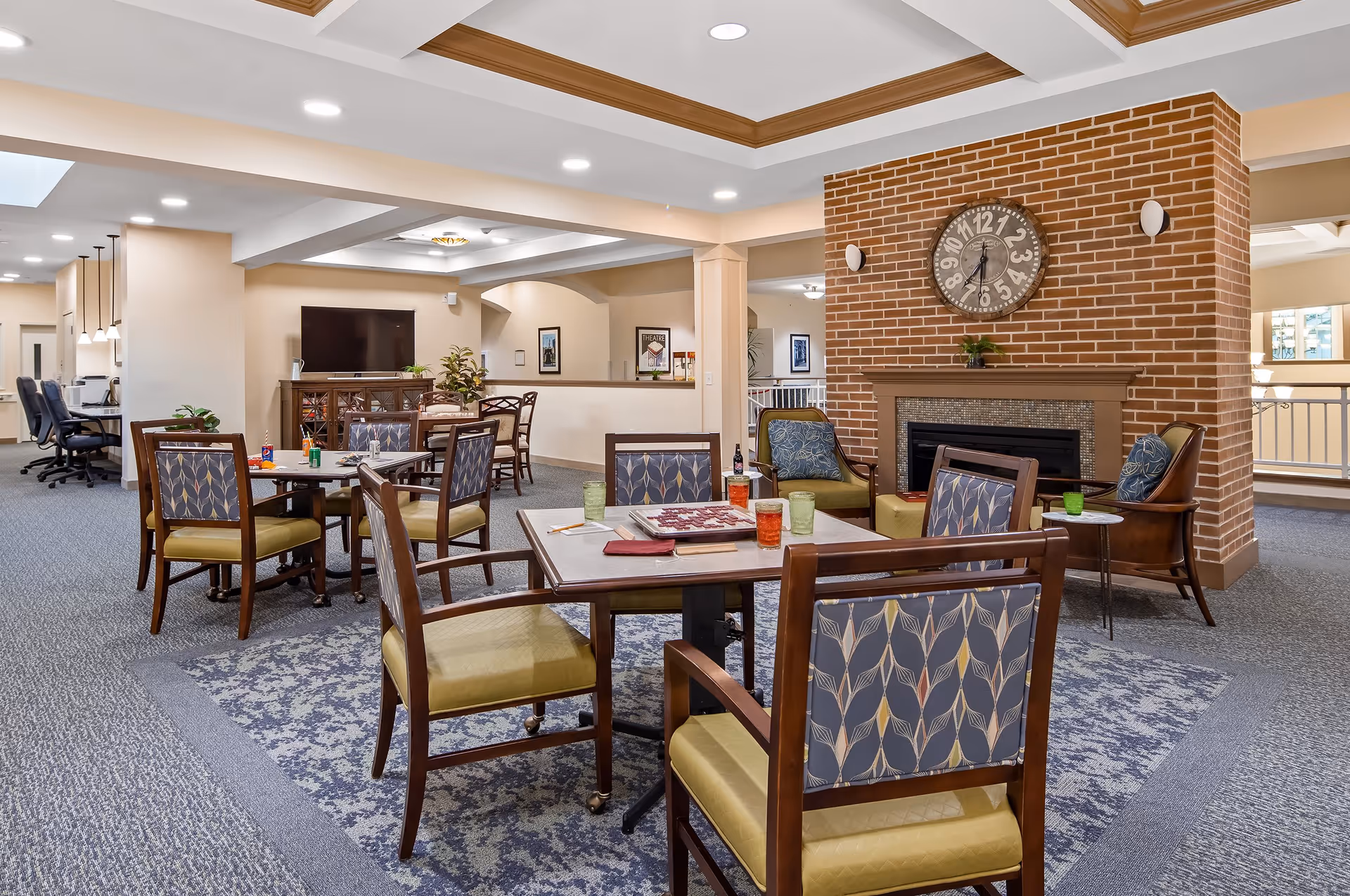 A spacious common area in a senior living facility featuring multiple tables with chairs arranged around them. The tables have board games and drinks on them. There is a brick fireplace with a large clock above it, and comfortable armchairs nearby. The room has carpeted floors, recessed lighting, and a television mounted on a cabinet in the background.