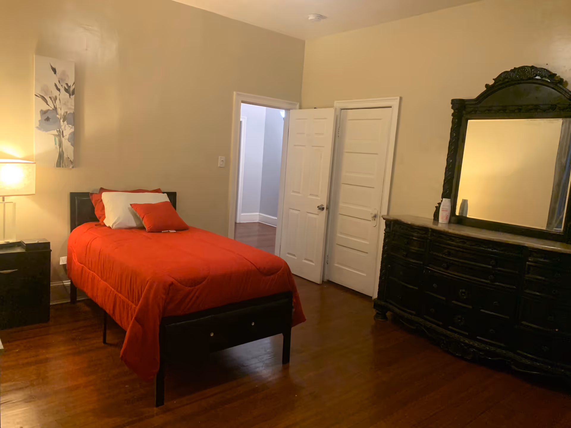 A furnished bedroom with a red quilted bed, nightstand, lamp, dresser, and mirror.