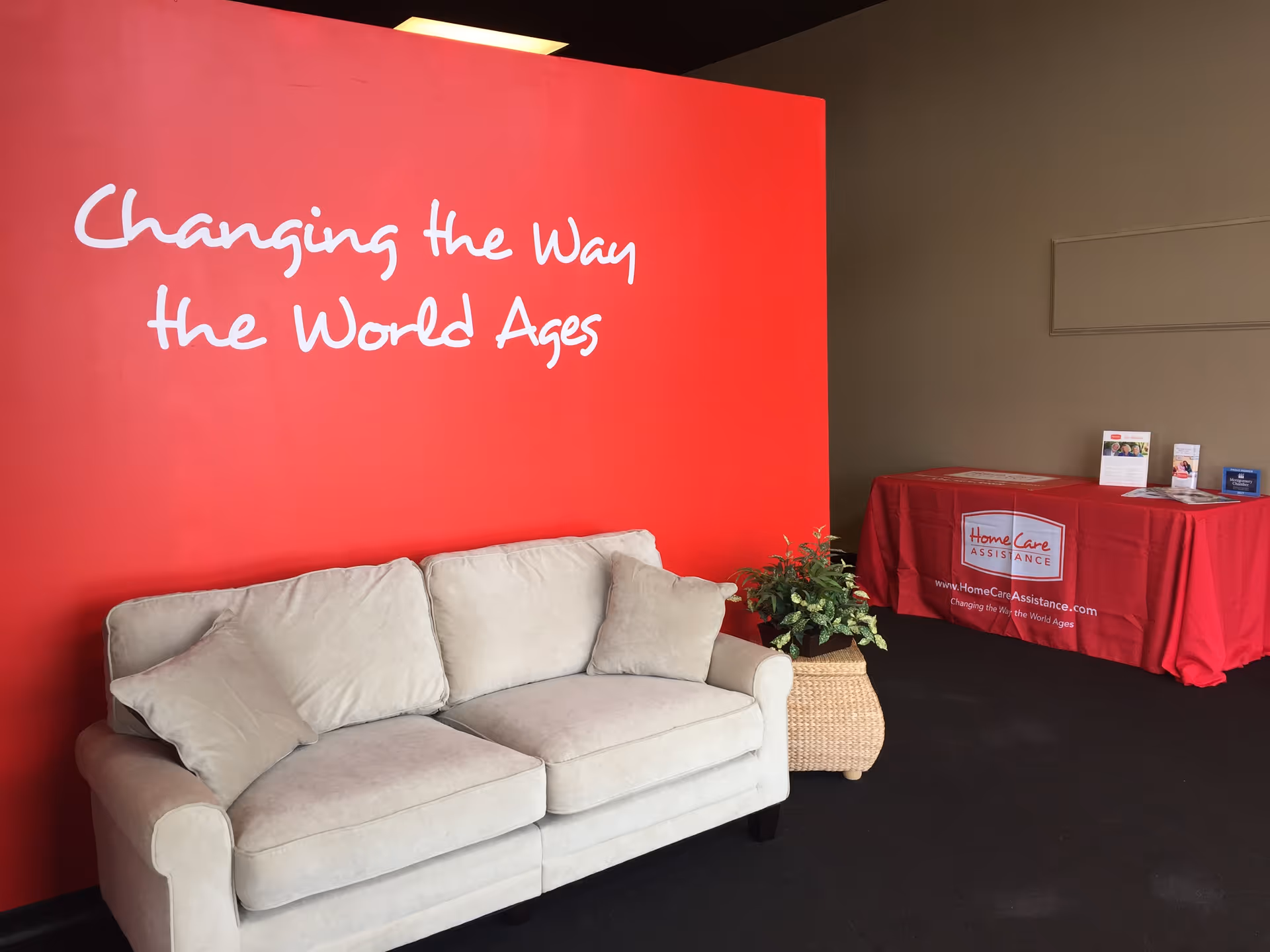 Red wall with inspirational slogan, beige sofa, potted plant, and branded table in lobby.