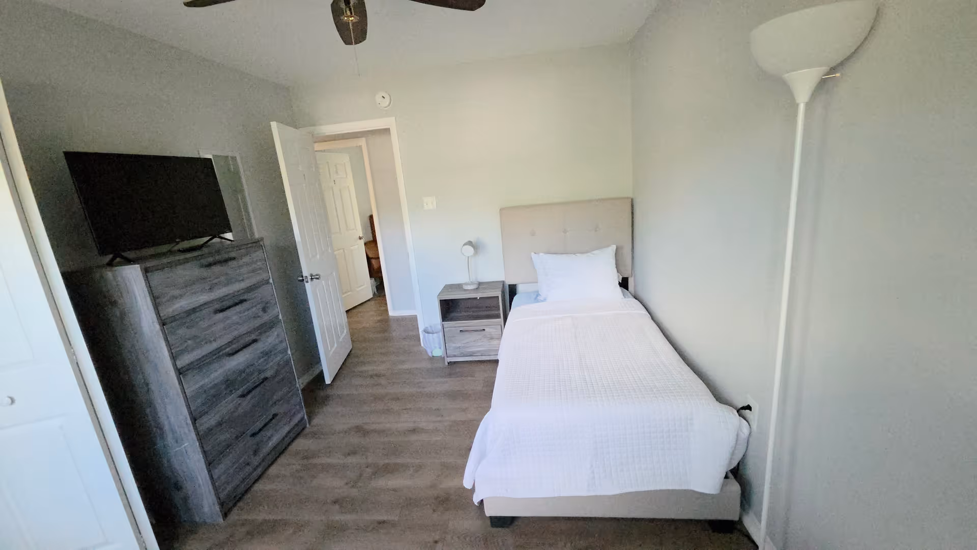 A small furnished bedroom with a single bed, dresser, nightstand, TV, and doorway.