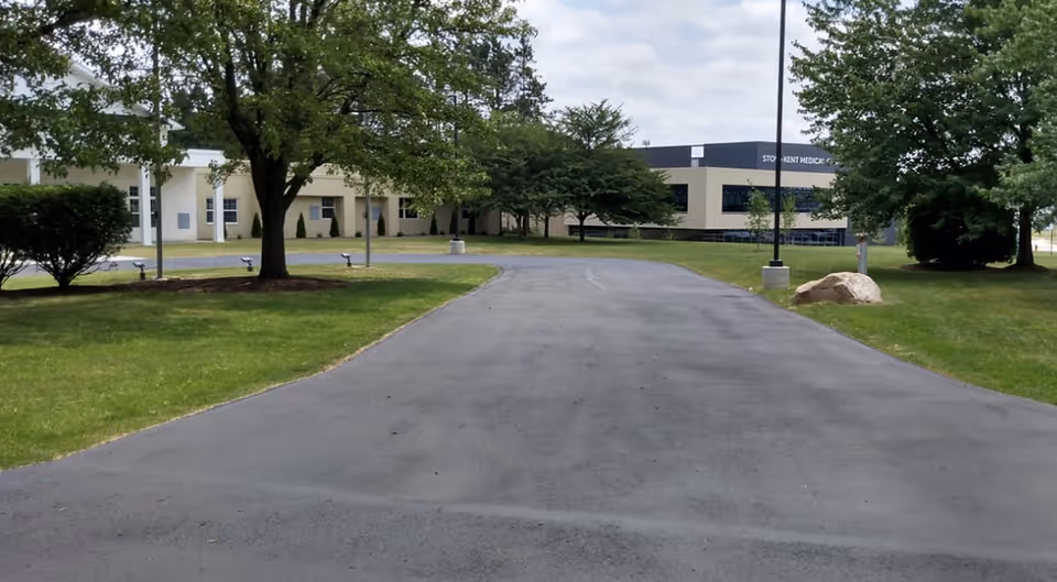 A paved driveway leads to the front of a low-rise beige senior living building surrounded by lawns and trees.