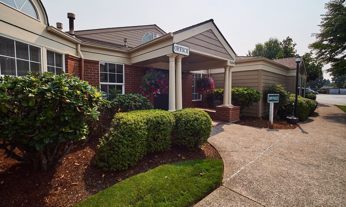 Brick senior living facility entrance with columns, hanging baskets, manicured shrubs, and a curved walkway.