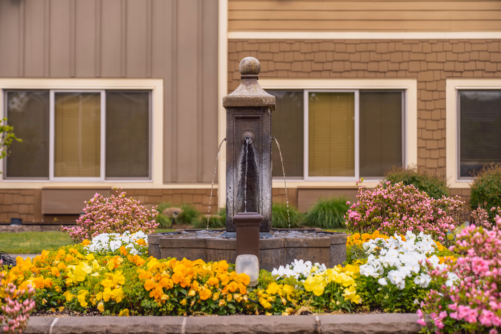 Stone fountain surrounded by colorful flowers in front of a beige senior living building.