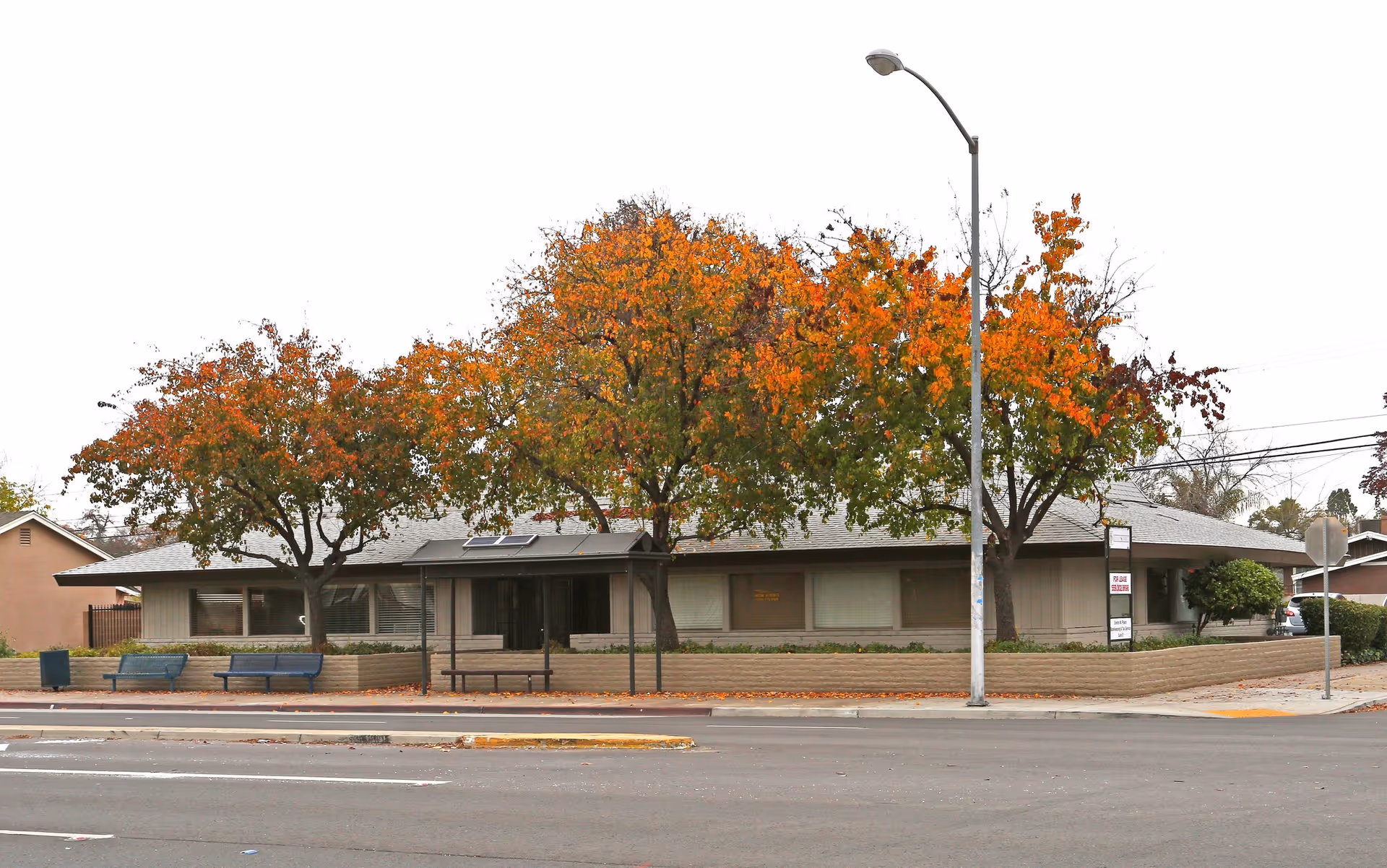 Single-story facility with brown brick exterior, street, benches, and colorful autumn trees.