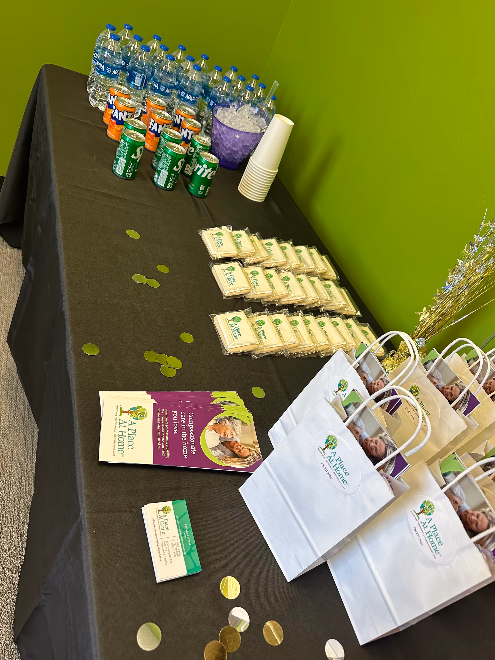 Refreshment table with bottled water, soda, bags, brochures, and gift bags against a bright green wall.