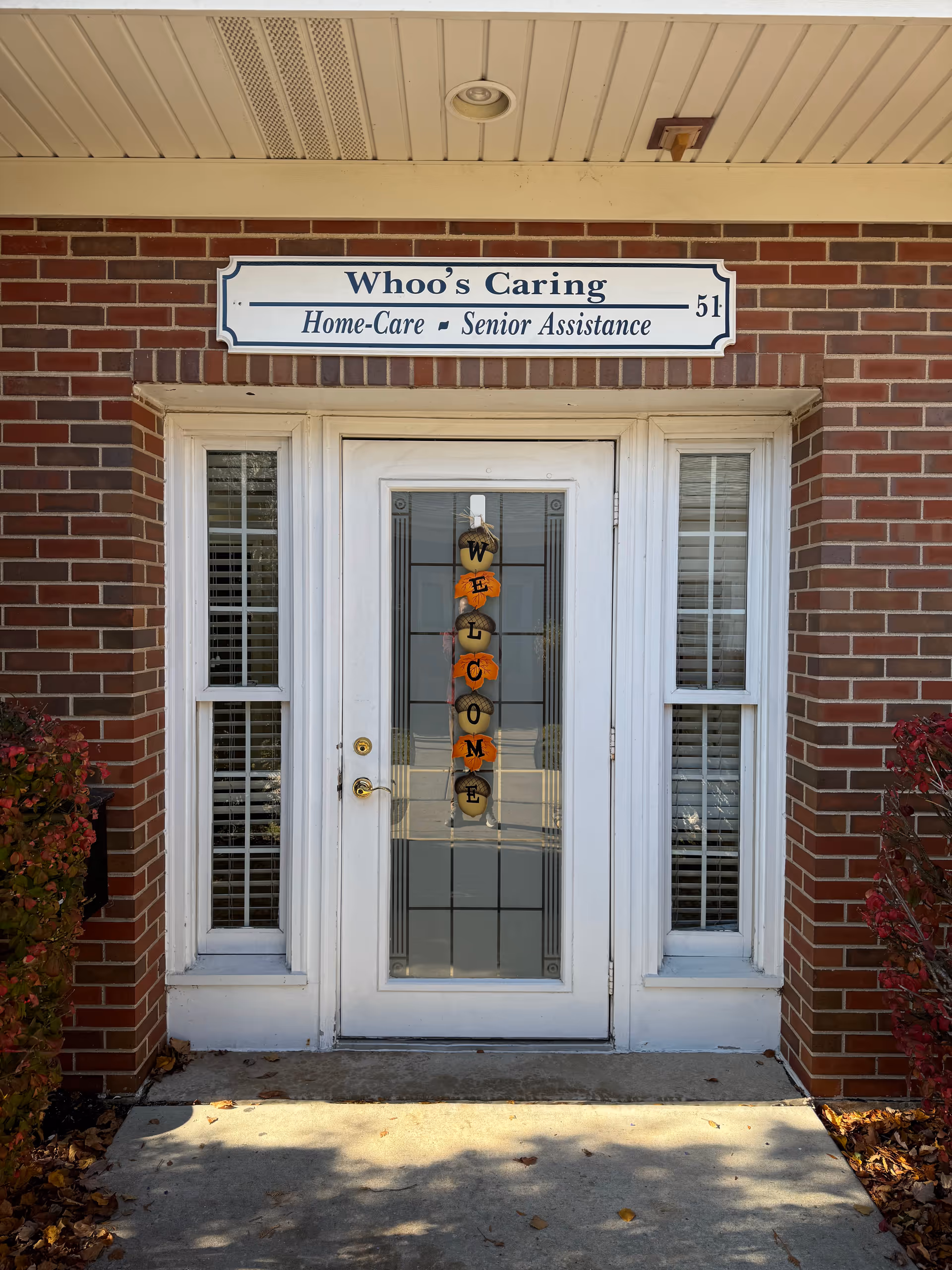 Brick building entrance with white door, Whoo's Caring sign, and pumpkins spelling WELCOME on glass