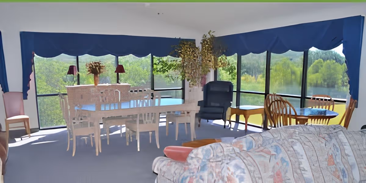 Communal lounge and dining area with tables, chairs, a sofa, and large windows overlooking trees and a pond.