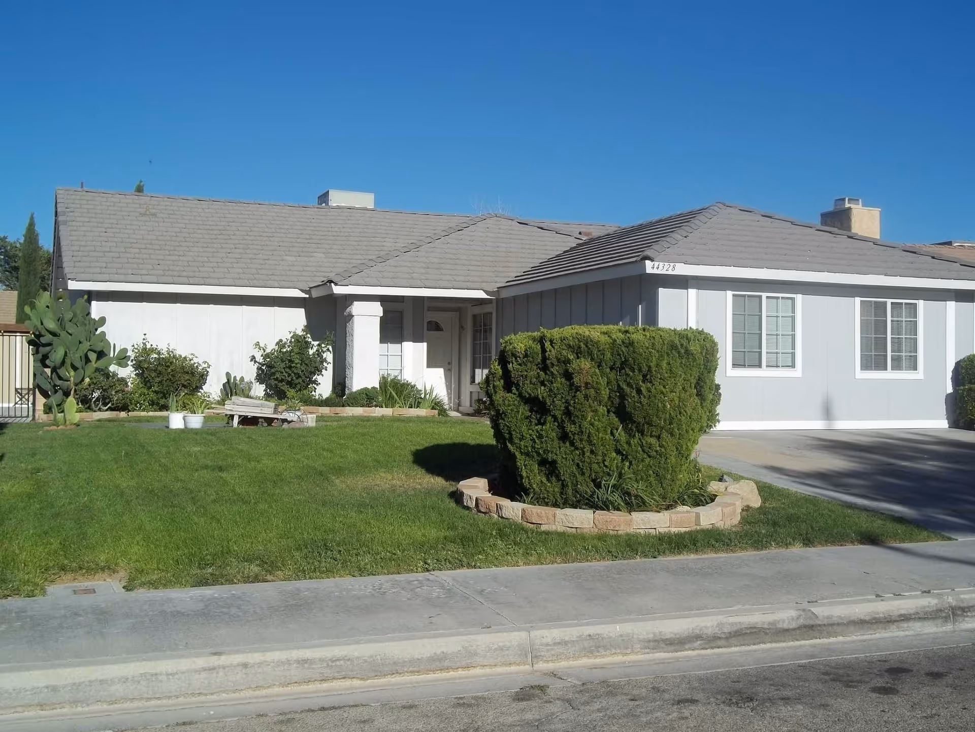 Single-story residential building with a gray roof and white exterior walls, surrounded by a well-maintained green lawn and trimmed bushes under a clear blue sky.