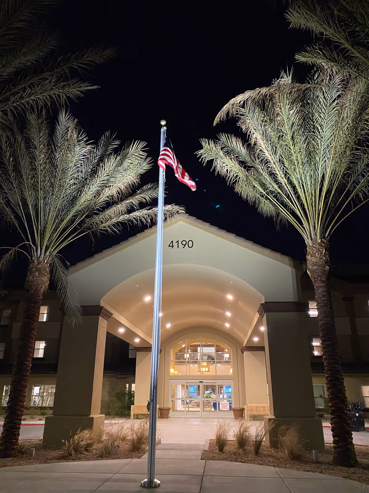 Night exterior of a retirement community entrance with flagpole and palm trees.