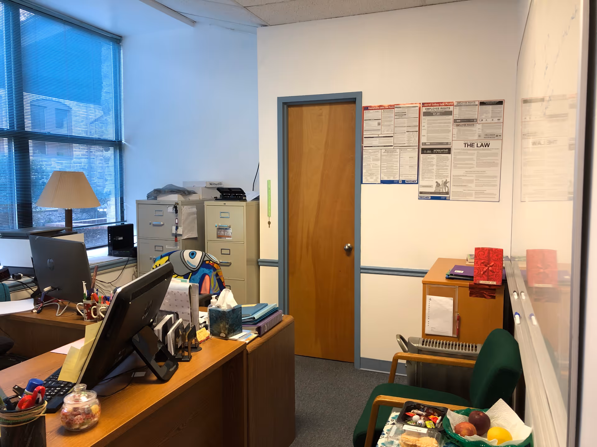 Office workspace with desks, computers, filing cabinets, and wall notices.