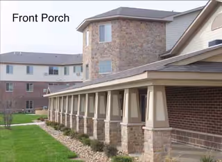 Exterior view of a senior living facility showing a long covered front porch with stone pillars and a multi-story building in the background. The building has a combination of brick and stone exterior walls and several windows.