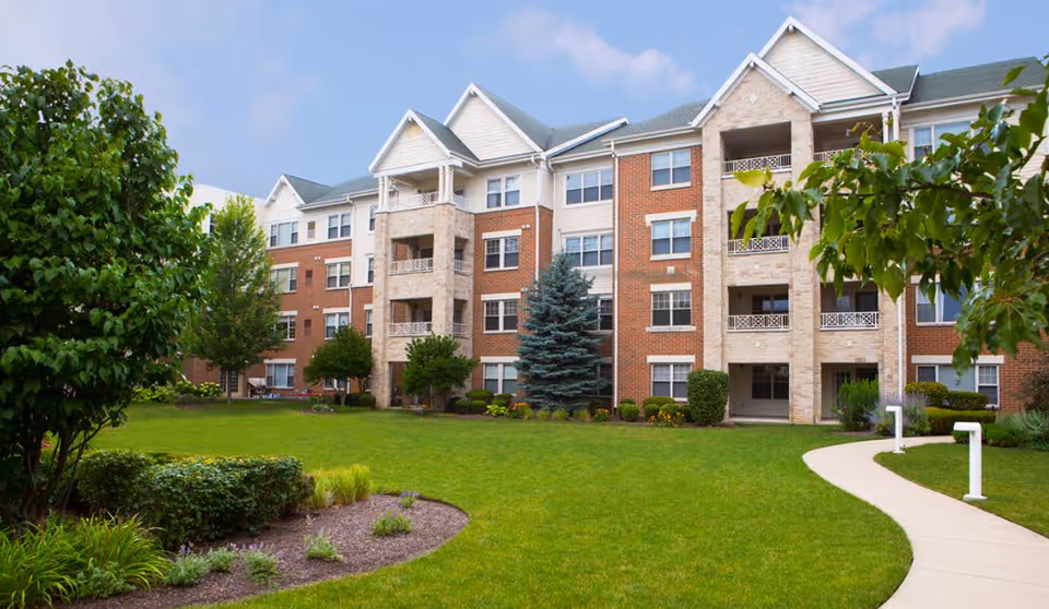 Exterior view of a multi-story brick and stone senior living building with landscaped lawn and curved walkway.
