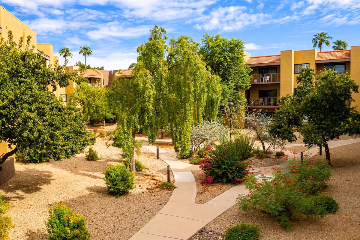 Sunlit courtyard with paved walkways, desert landscaping, trees and shrubs between beige apartment buildings under a blue sky.