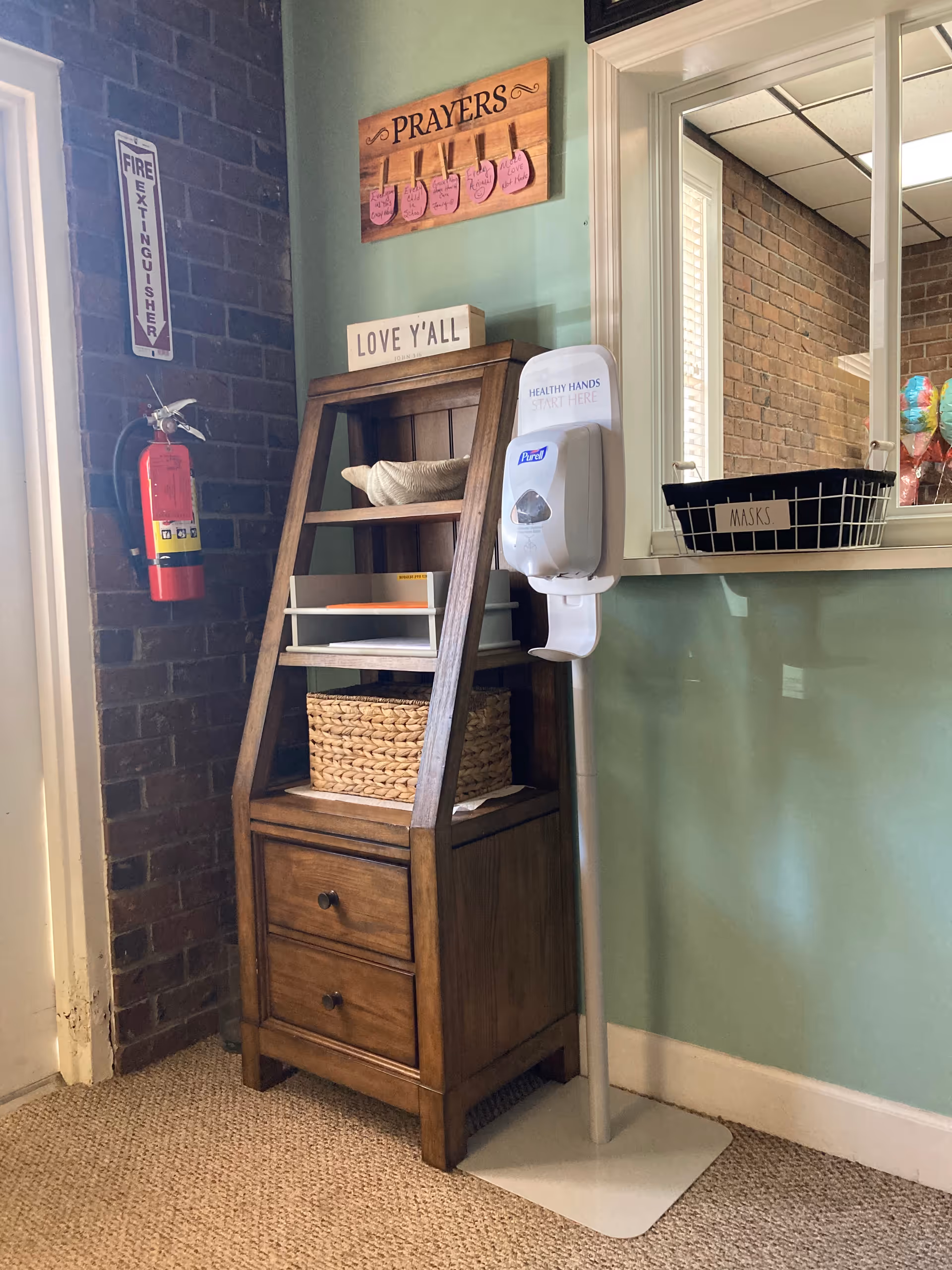 Indoor facility corner with wooden shelf, hand sanitizer, prayer signs, and a masks basket.