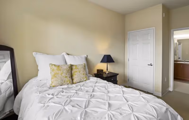 Bedroom with a neatly made bed, pillows, nightstand, lamp, and doorway to a bathroom.