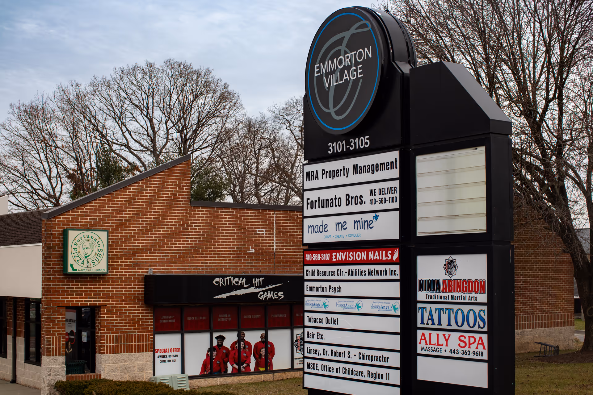 Brick commercial building with a tall signboard listing businesses and bare trees behind.
