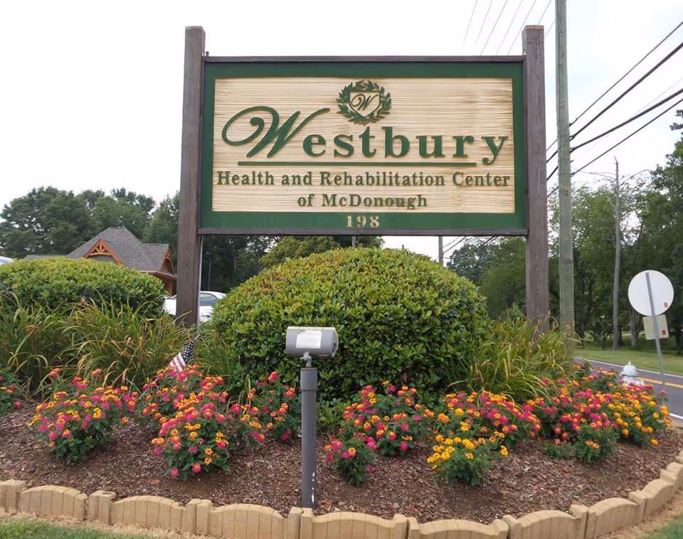 A large wooden sign reading Westbury Health and Rehabilitation Center of McDonough, surrounded by flowers.