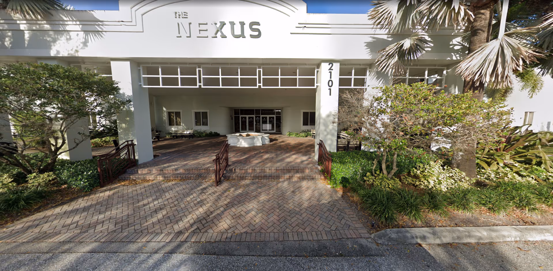 Brick-paved walkway leads to a building entrance labeled The Nexus, with greenery and palm trees.