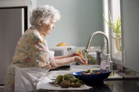 Elderly woman wearing an apron chops vegetables at a kitchen sink.