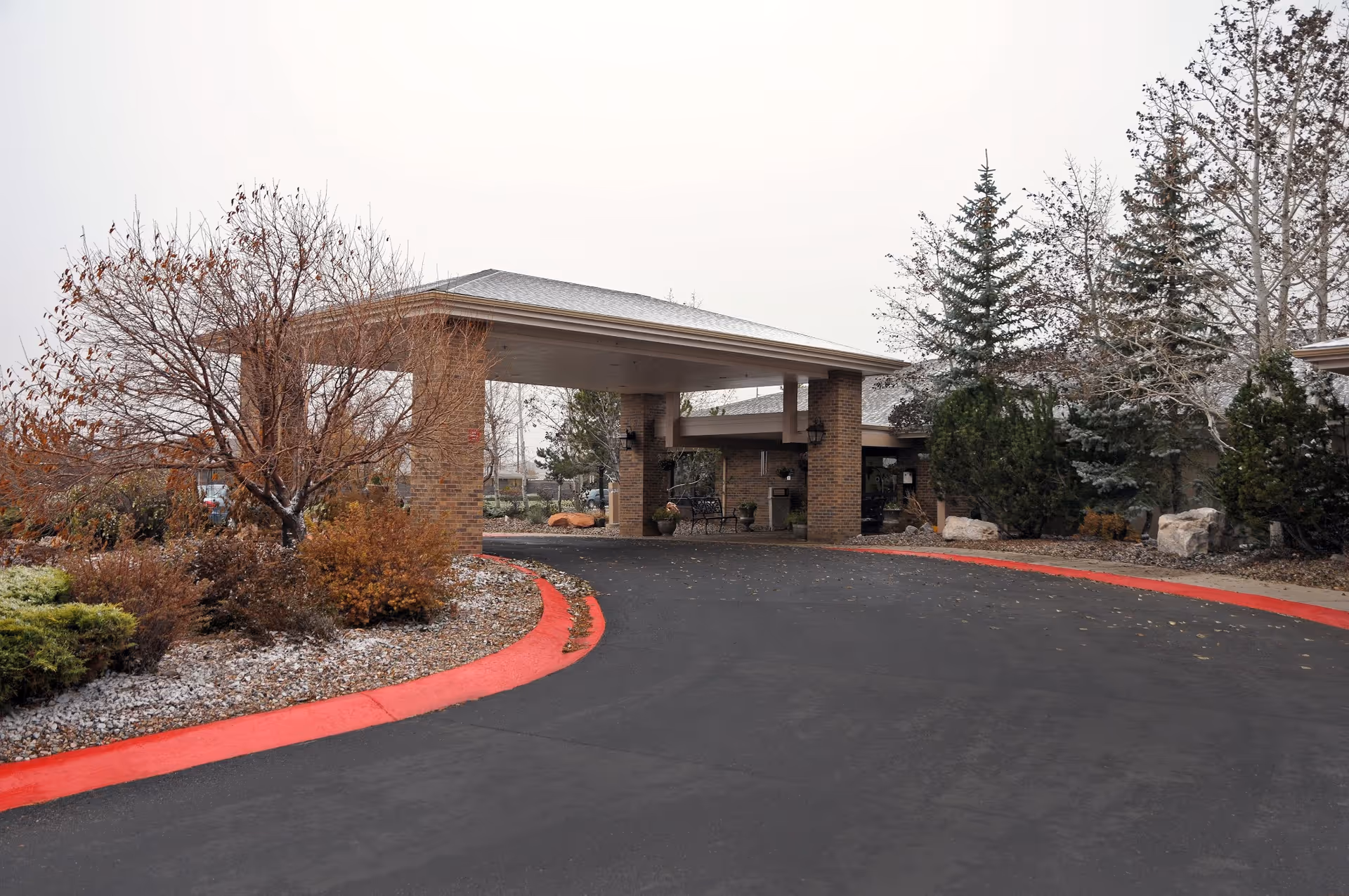 Front entrance of a brick senior living facility with a covered porte-cochere, curved driveway, and landscaping.