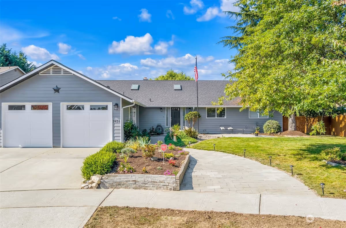 A single-story blue-gray home with two-car garage, landscaped front yard, and American flag.