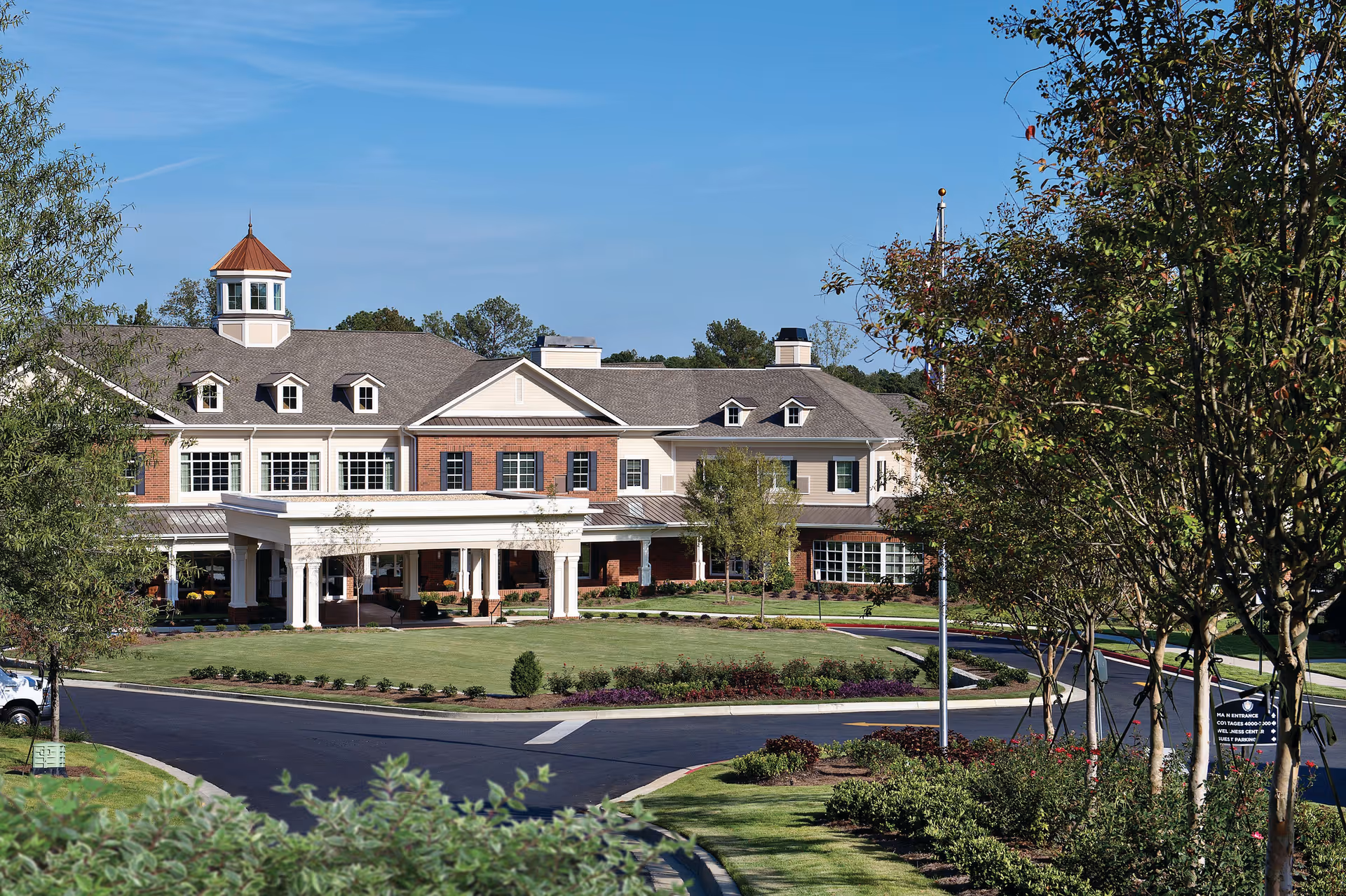 Exterior view of Sterling Estates of East Cobb Retirement Community building with a covered entrance, manicured lawn, trees, and clear blue sky.