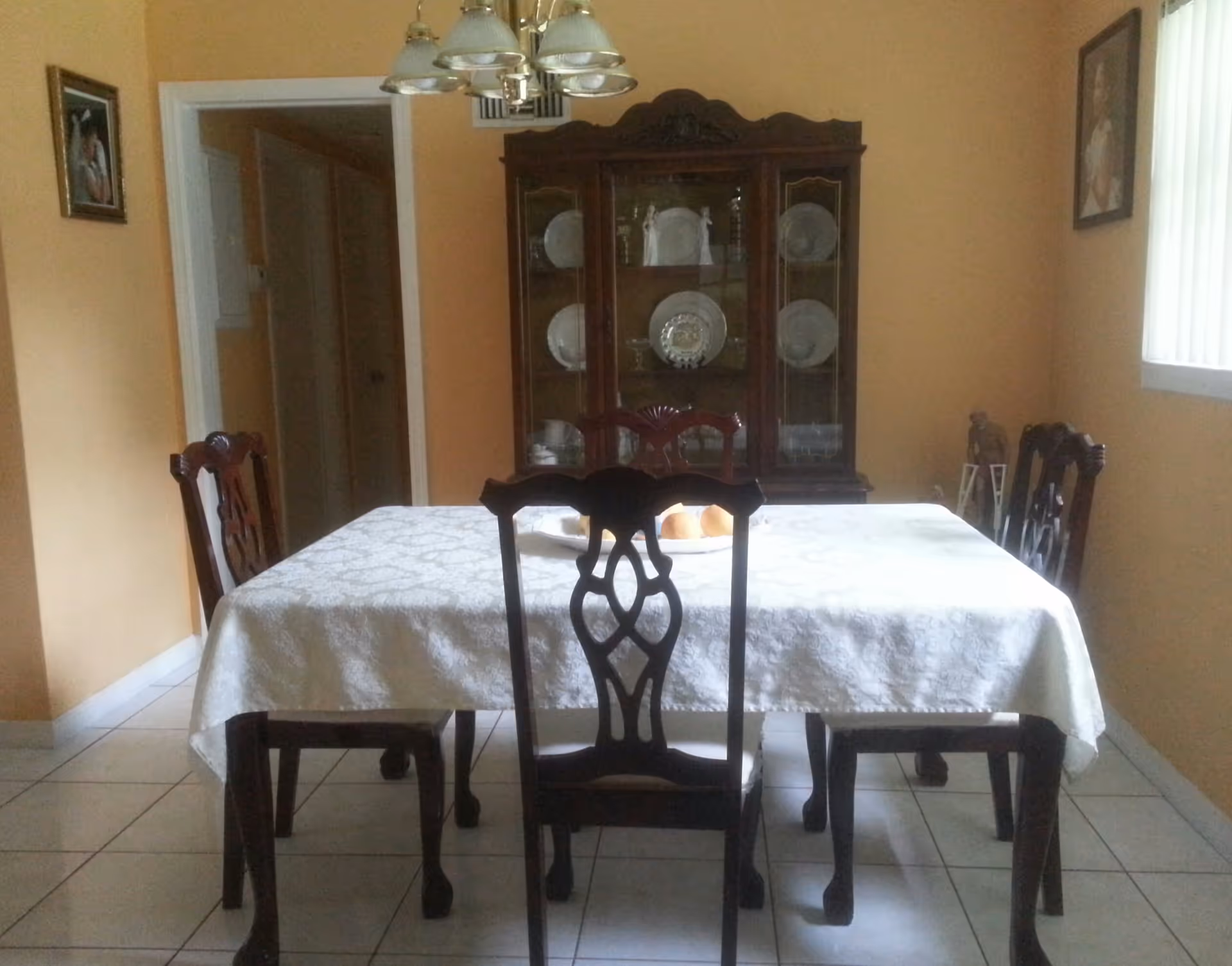 A dining room with a rectangular table covered with a white patterned tablecloth and surrounded by six wooden chairs. A wooden china cabinet with glass doors displaying plates and glassware is against the back wall. The walls are painted a warm beige color, and there are framed pictures on the walls. A window with vertical blinds allows natural light into the room, and a ceiling light fixture with multiple lamps hangs above the table.