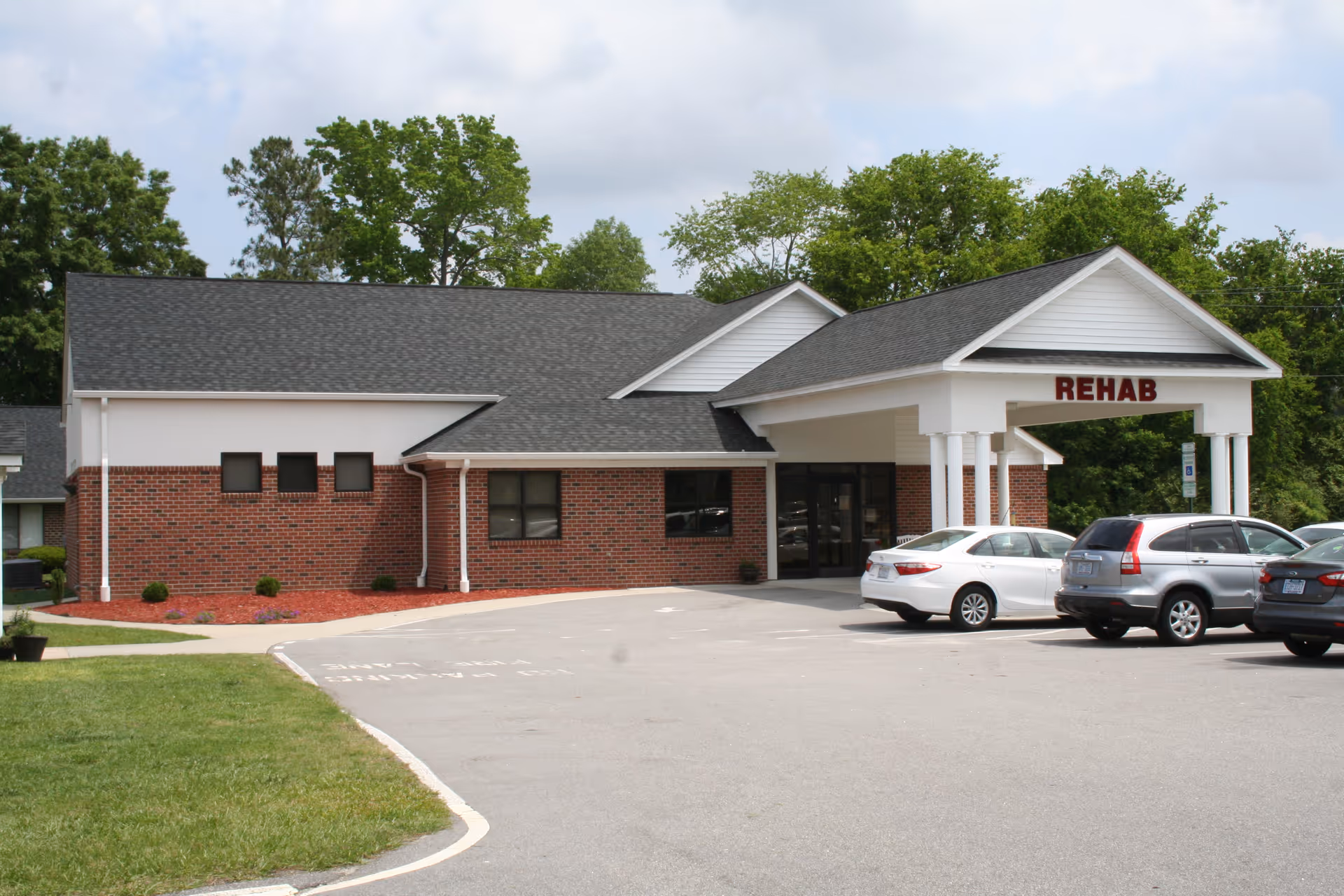Single-story brick rehab facility with a covered entrance and parked cars in front.