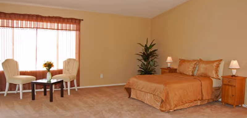 A furnished senior living bedroom with a bed, nightstands, lamps, a plant, and seating by window.