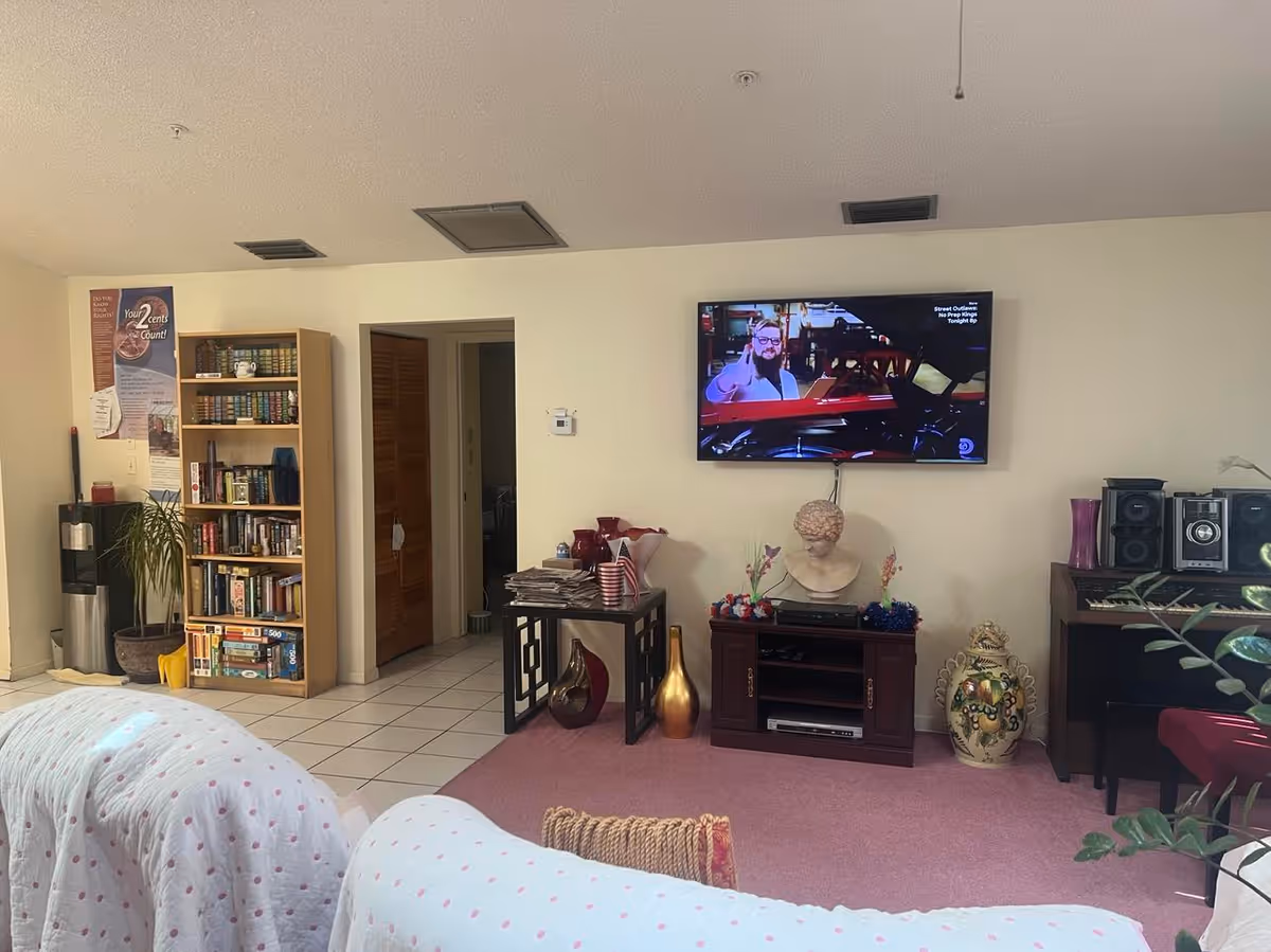 A cozy living room area in a senior living facility with a wall-mounted TV showing a man giving a thumbs up. Below the TV is a wooden cabinet with decorative items including a bust sculpture and vases. To the left, there is a bookshelf filled with books and a water dispenser next to a potted plant. The room has tiled flooring near the bookshelf and carpeted flooring in the seating area with white furniture covered in white fabric with pink polka dots.