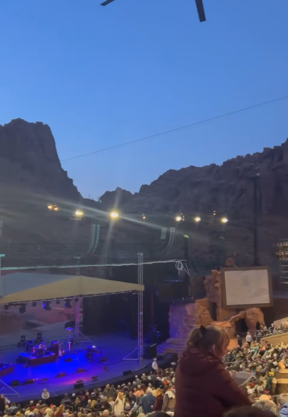 An outdoor concert venue with a stage, audience seating, and rugged rock backdrop.