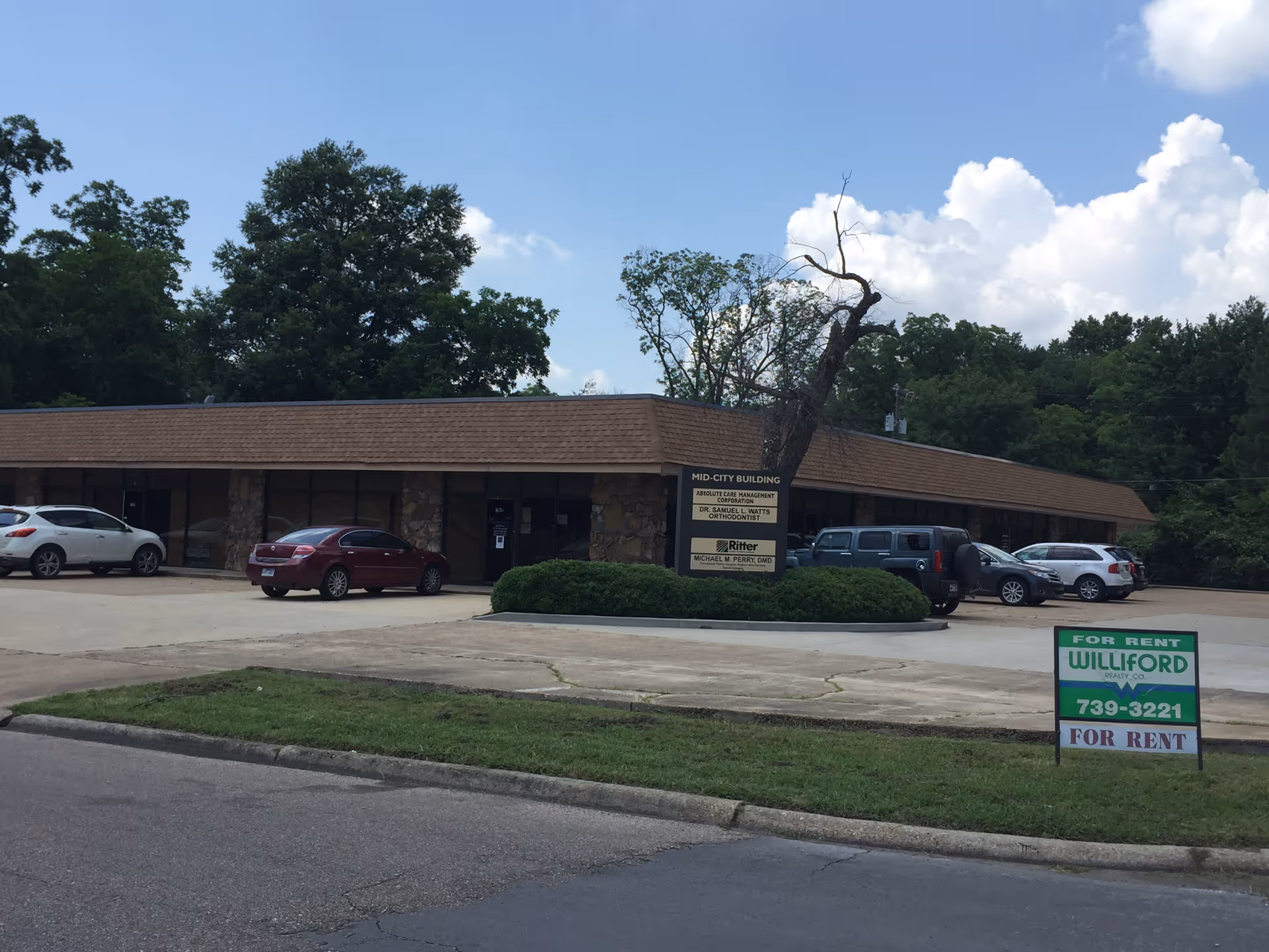 Single-story commercial building with a parking lot and trees behind, sign near driveway.