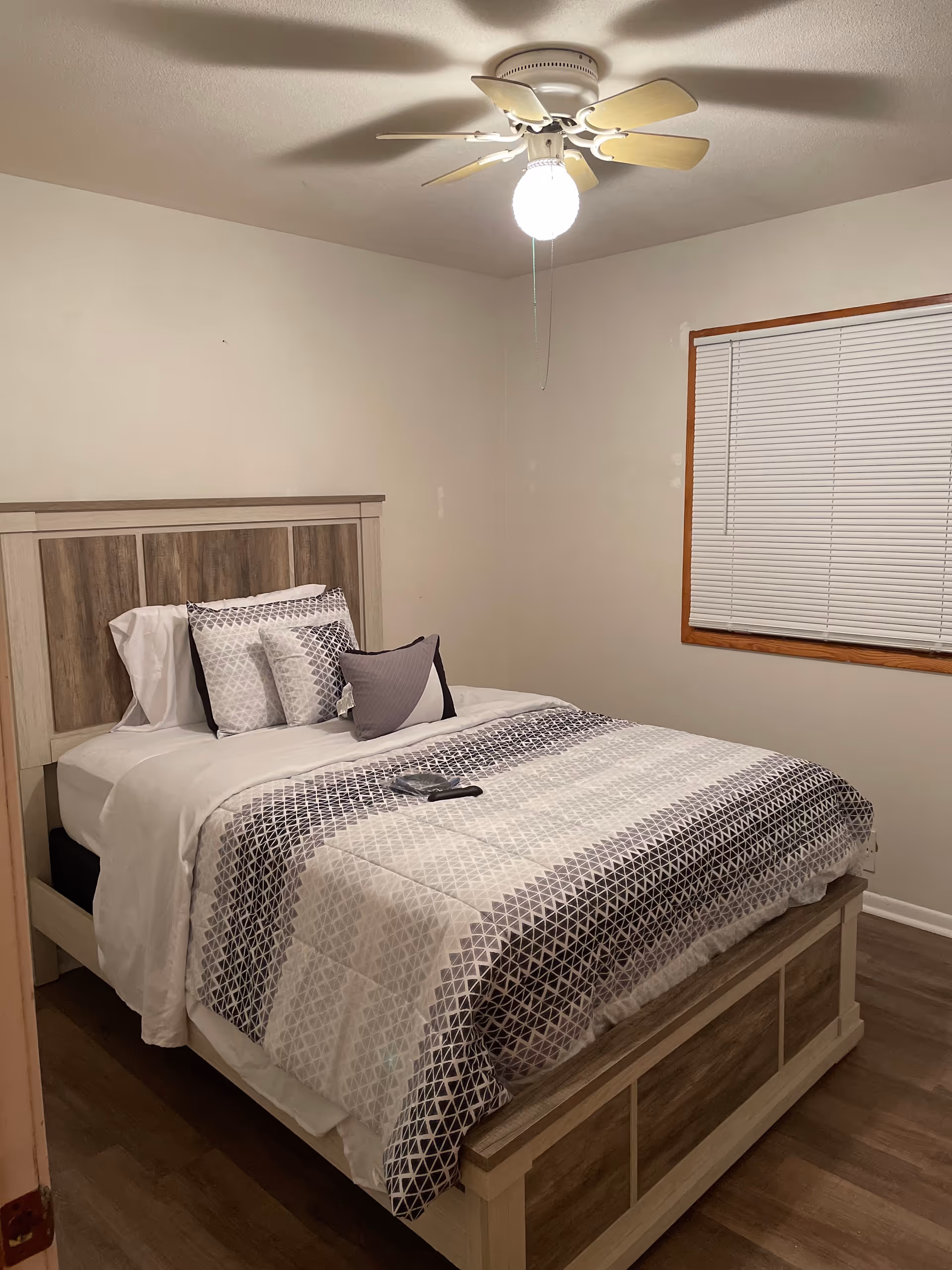 A neatly made bed in a cozy bedroom with a ceiling fan and blinds.