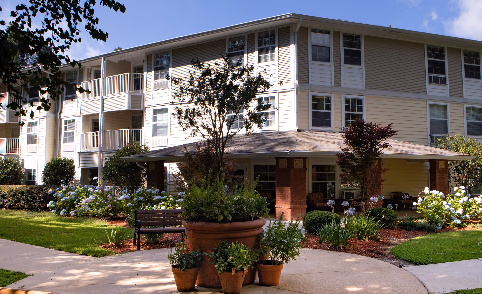 Front exterior of a multi-story senior living building featuring a covered entrance, balconies, a landscaped courtyard, benches, and potted plants.