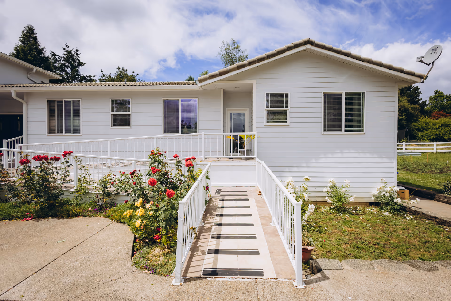 White single-story facility exterior with a wheelchair ramp, garden, and roses