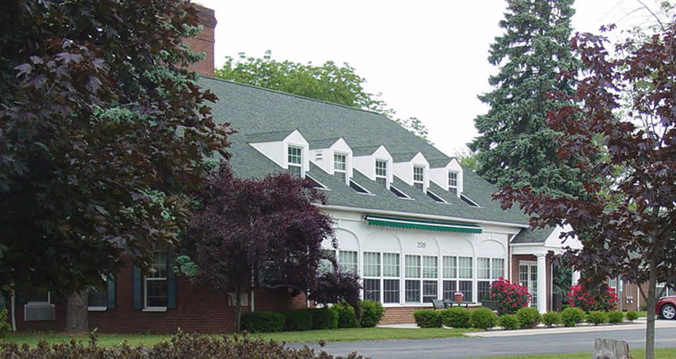 Front exterior of a brick senior living facility with dormer windows, a green awning, large front windows and landscaped trees and shrubs.