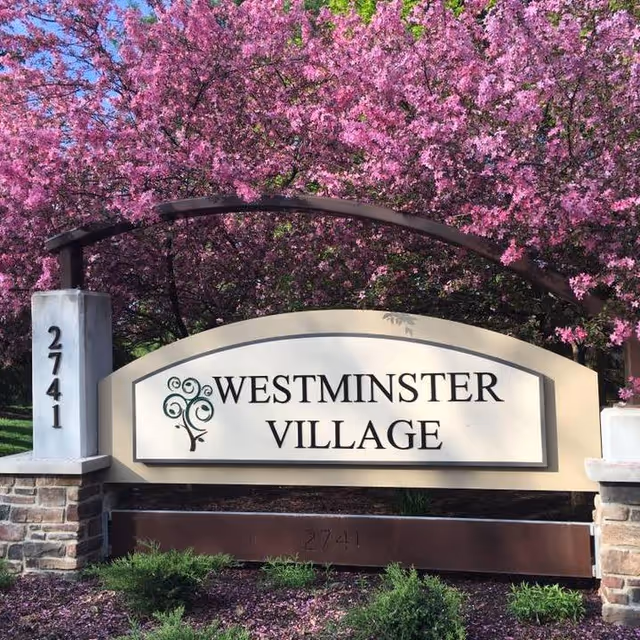 Entrance sign reads Westminster Village with flowering pink trees behind it.