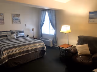 A cozy bedroom featuring a large bed with striped bedding and pillows, a window with blue curtains, a floor lamp next to a small wooden side table, and a dark brown leather armchair with a light-colored pillow.