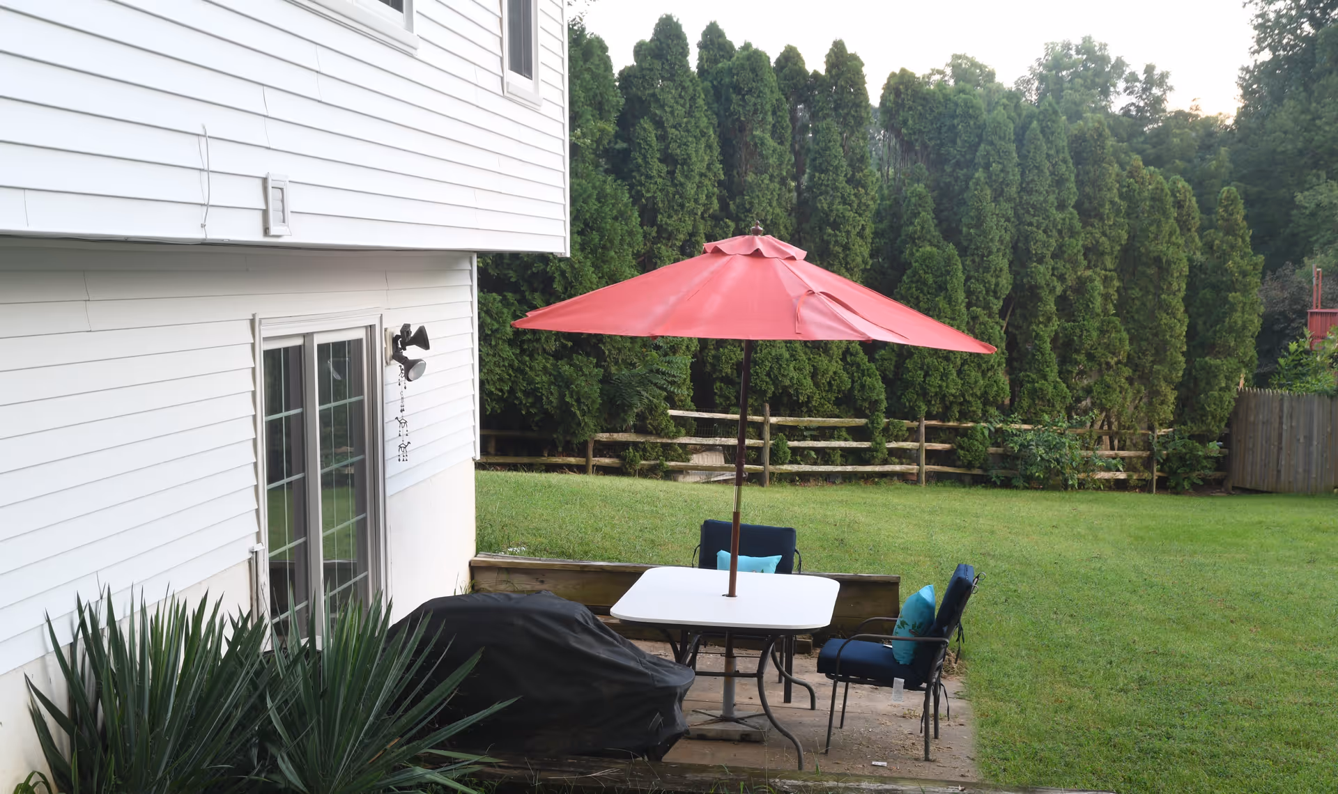 Outdoor patio area with a white table, two blue cushioned chairs, and a red umbrella. The patio is adjacent to a white house with sliding glass doors. There is a covered grill and green grass lawn with tall evergreen trees and a wooden fence in the background.