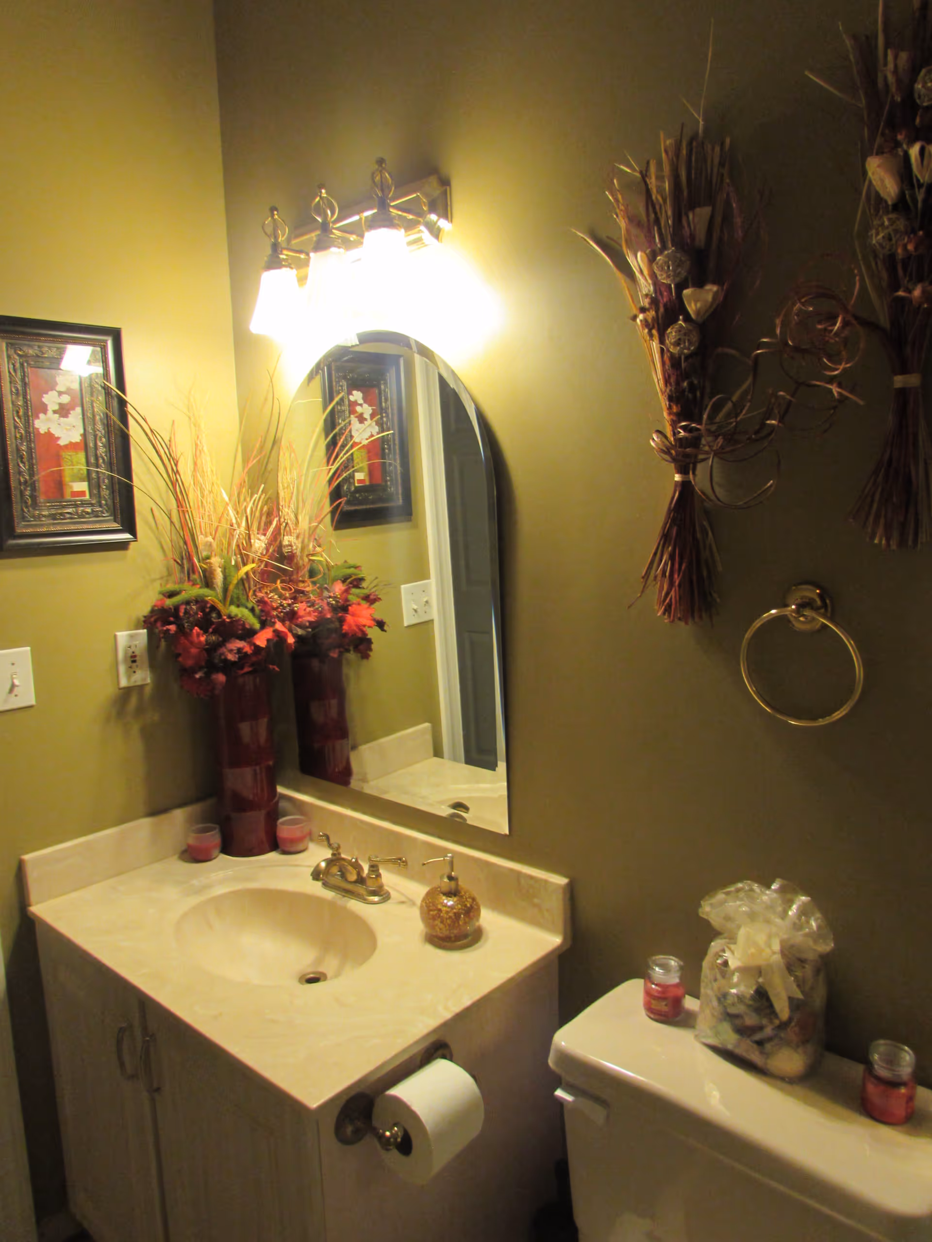 A bathroom with a beige countertop sink, a wall-mounted mirror with three light fixtures above it, a toilet with decorative items on top, and wall decorations including a framed picture and dried floral arrangements.