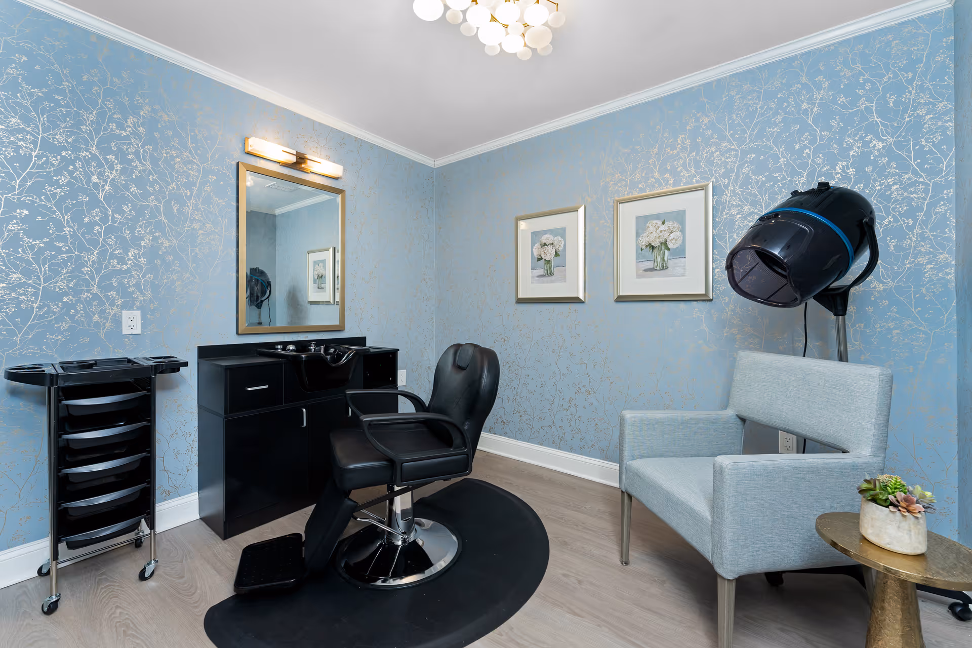 A small salon room with a black salon chair in front of a black cabinet with a sink and a mirror above it. There is a black rolling cart with trays on the left side and a light blue armchair with a hair dryer hood on the right side. The walls are light blue with a subtle floral pattern, and two framed floral pictures hang on the wall. A modern light fixture is on the ceiling.