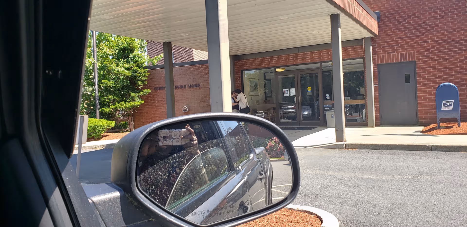 Car side mirror frames a building entrance with glass doors at Nevins Nursing & Rehab Center.