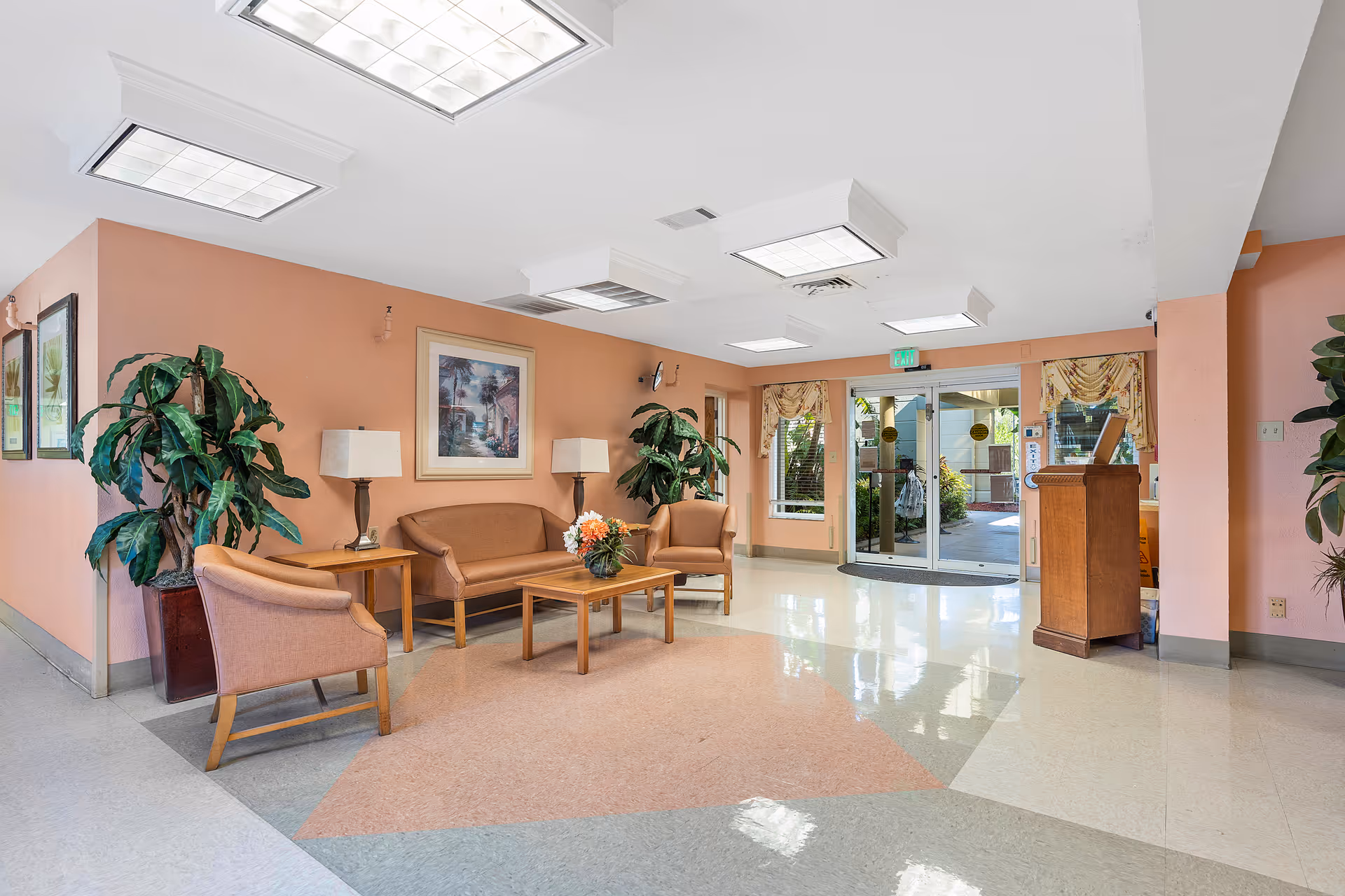 A bright and clean lobby area with peach-colored walls, featuring a seating arrangement with a sofa, two armchairs, a wooden coffee table with a flower arrangement, and two side tables with lamps. Large potted plants flank the seating area. The floor has a geometric pattern, and the ceiling has multiple square fluorescent light fixtures. Glass double doors lead outside, with windows on either side and floral curtains above.