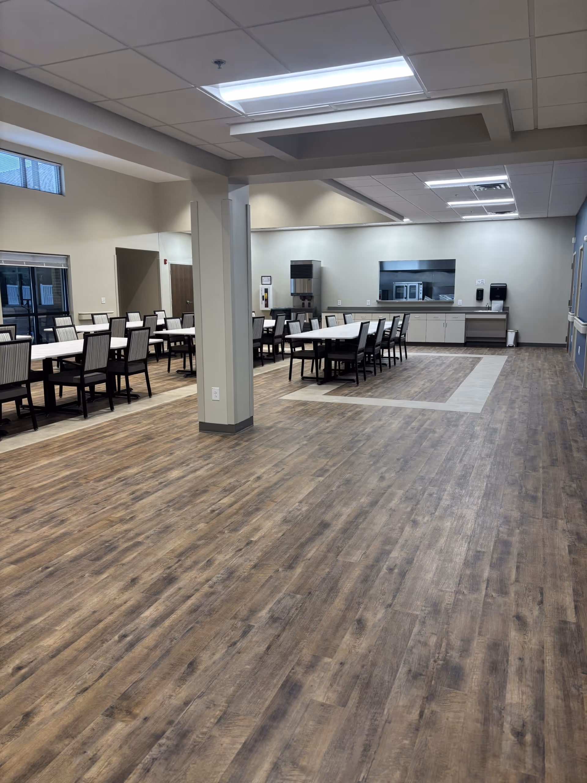 Spacious dining hall with rows of tables and chairs, kitchenette at the far end.