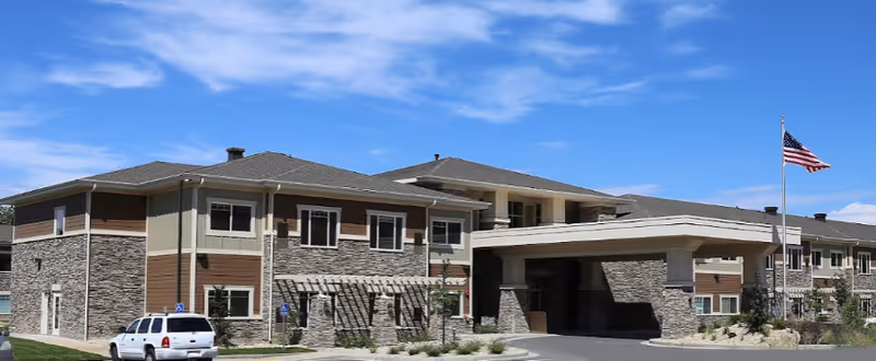 Exterior front view of a two-story senior living building with a covered entrance, parked car, and an American flag.
