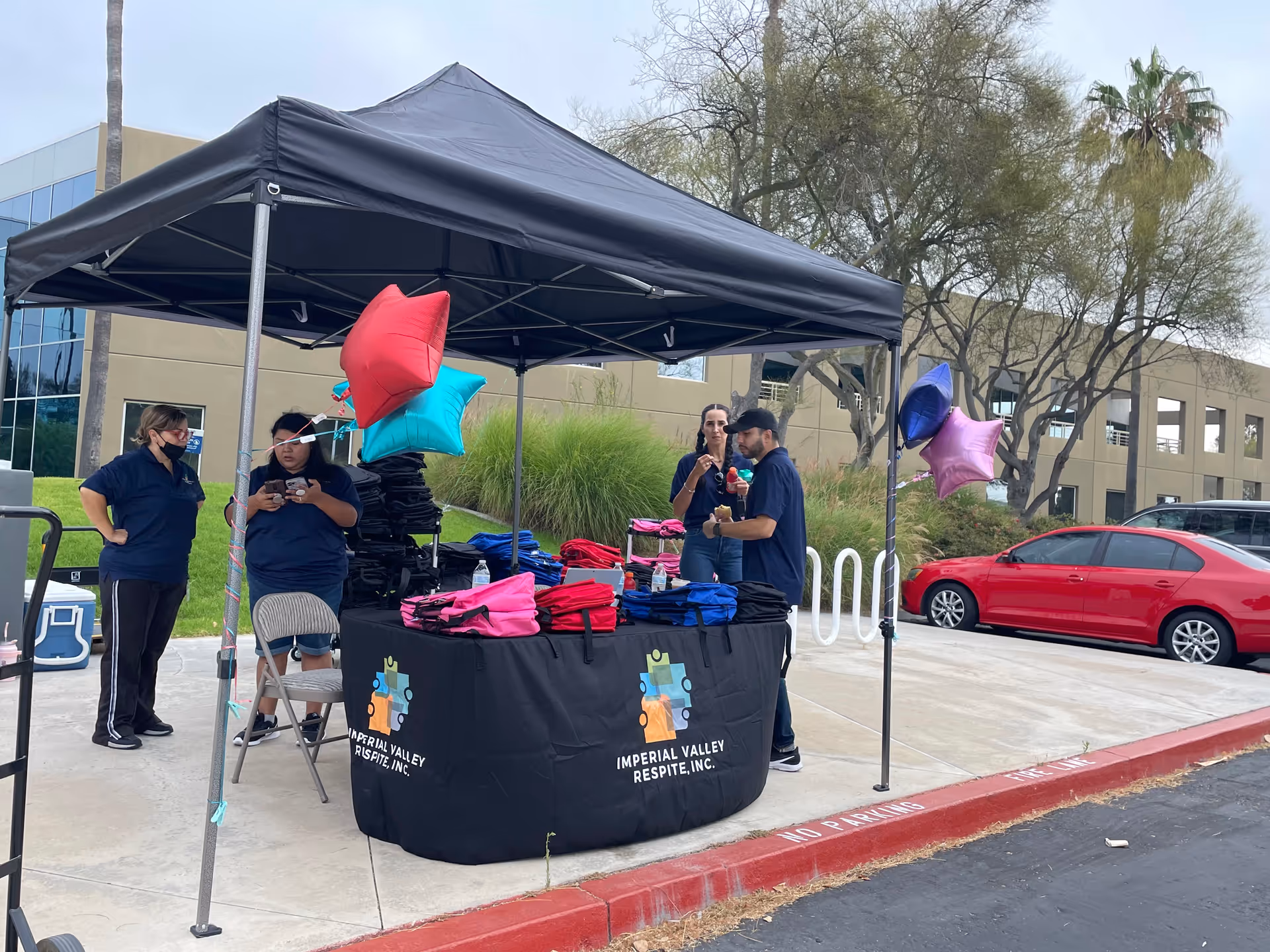 Outdoor tent setup with staff distributing items at a table; colorful balloons nearby.