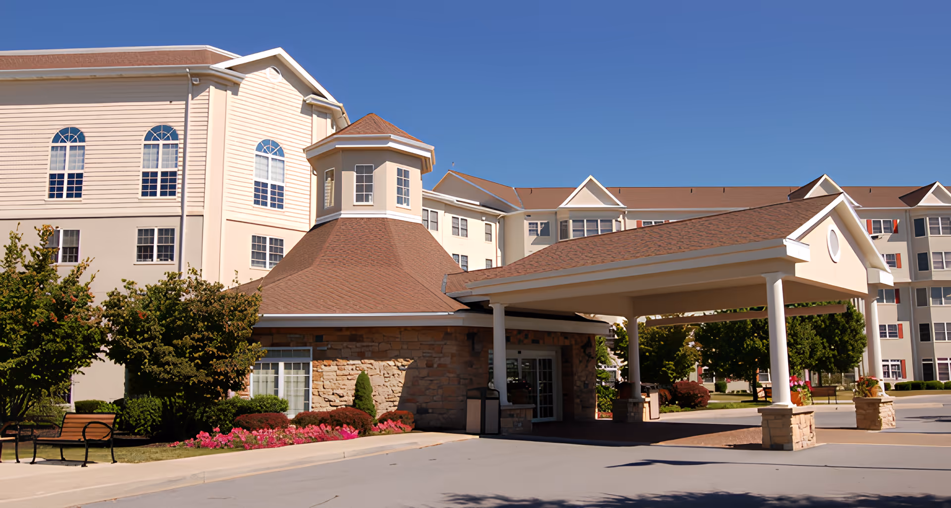 Exterior view of a senior living facility building with beige siding and a brown roof under a clear blue sky. The entrance features a covered driveway supported by white columns and stone bases. There are landscaped bushes, flowers, and benches near the entrance.
