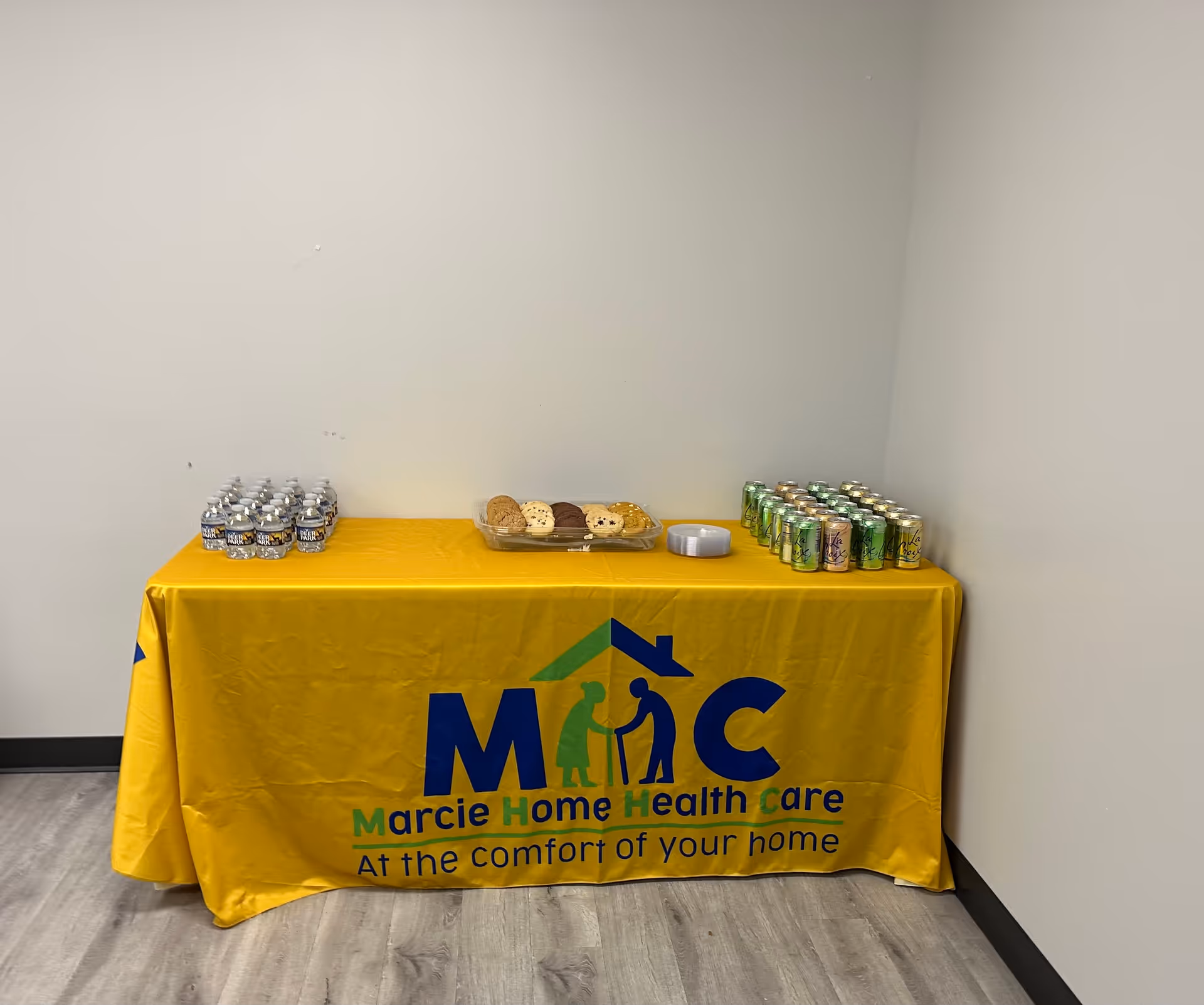 Yellow tablecloth-covered table with cookies and canned drinks against a plain wall.